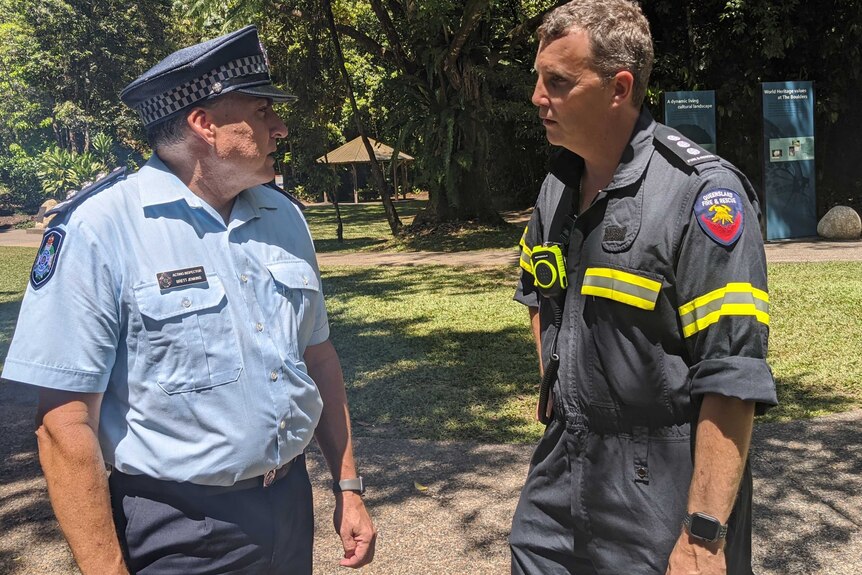 Police officer and fire officer stand in a park talking.
