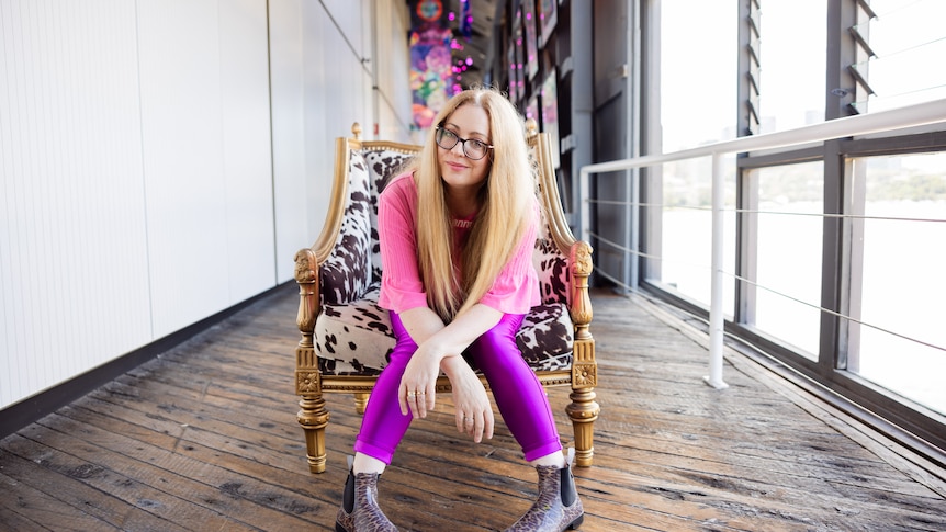 A white woman with blonde hair in her late 40s wears a bright pink shirt and purple leggings and sits in a cow hide chair.