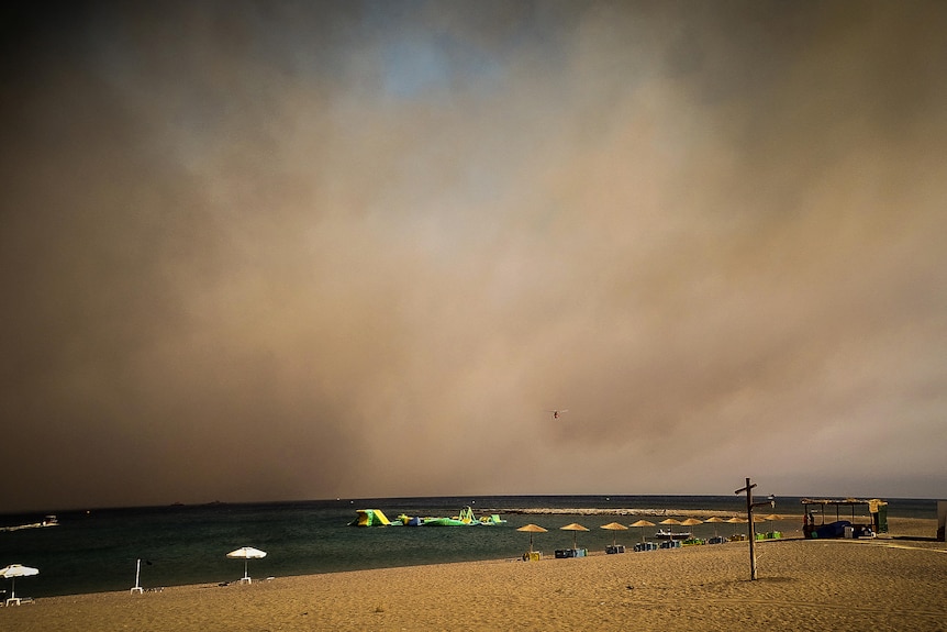 A beach on Rhodes is empty of people and a helicopter hovers above as wildfire fills the sky