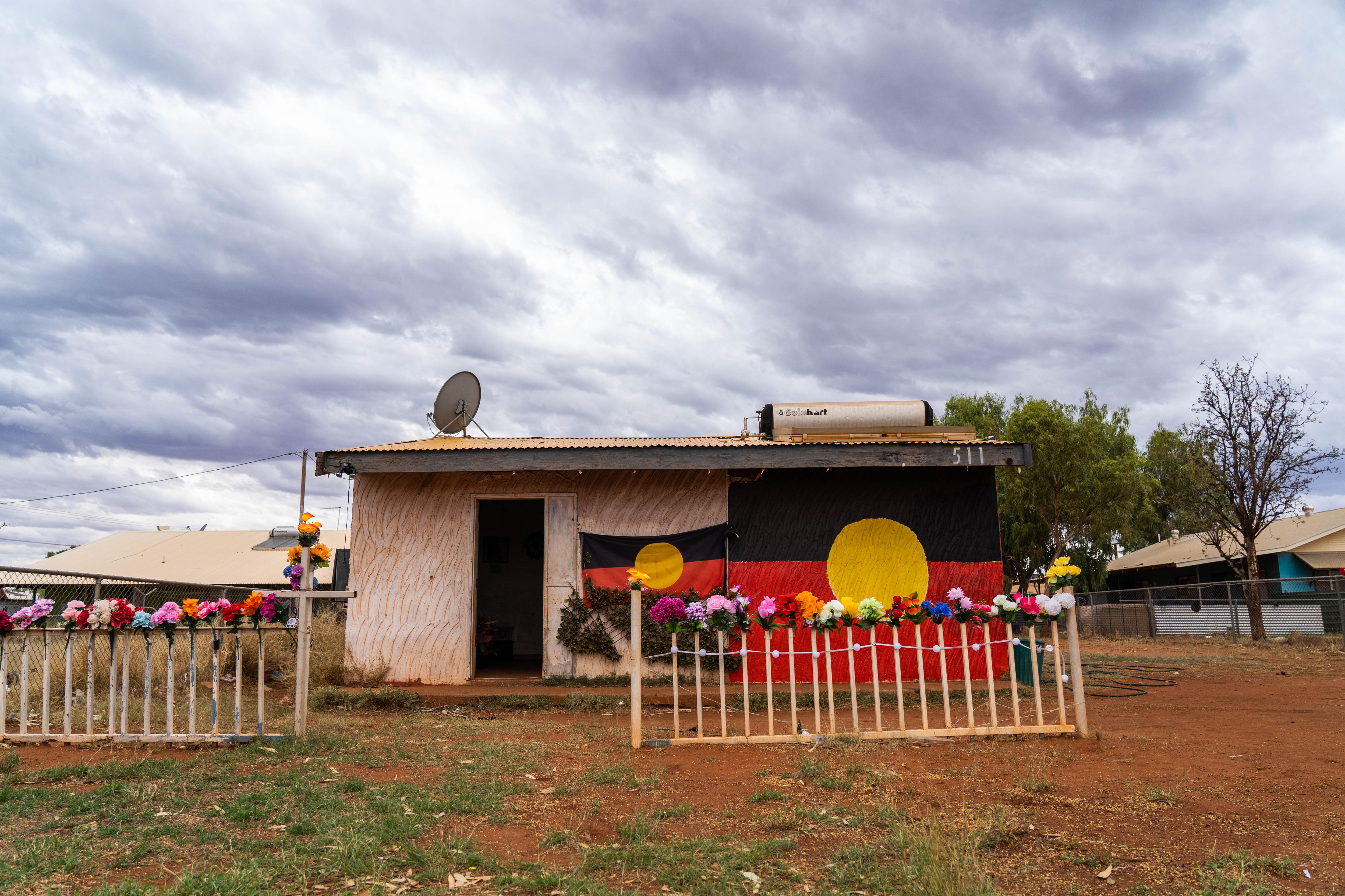 一个偏远社区的小型社区房屋，前面有两面原住民旗帜，前围栏两旁摆着鲜花