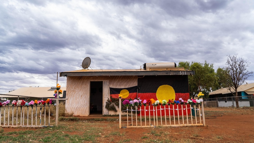 A small community house in a remote community with two Indigenous flags on the front, and flowers lined along its front fence