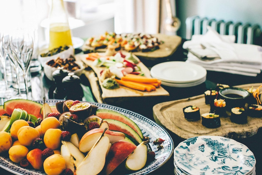 Table filled with fruit platter, sushi and other foods, wine glasses and plates.