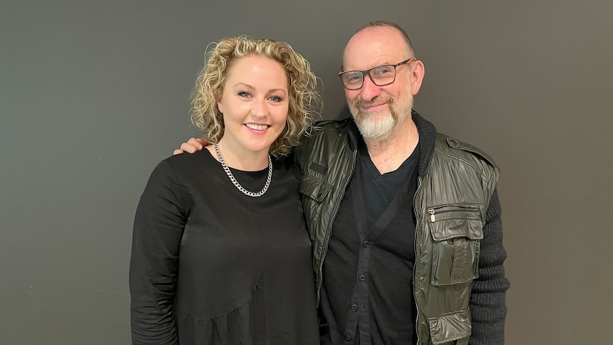 Zan Rowe and Colin Hay stand against a black wall, they are both wearing black clothes and smiling