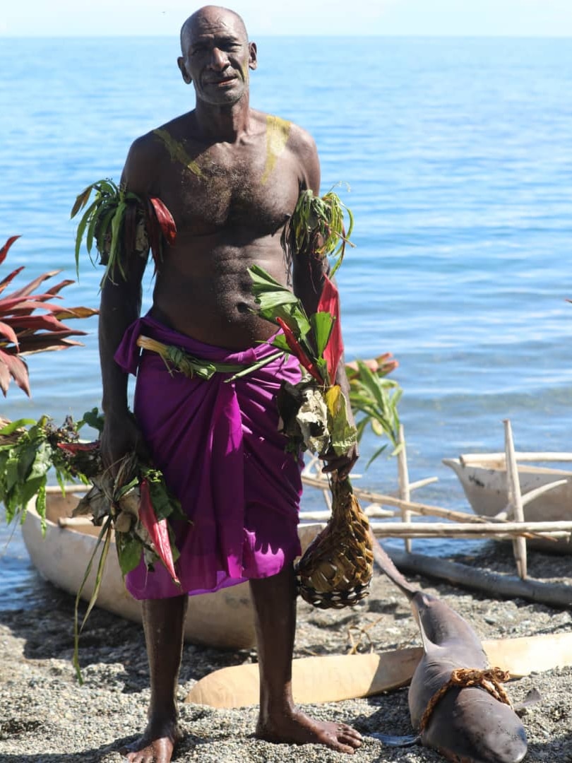 Man stands bare chest wearing purple sarong and shark by his feet.