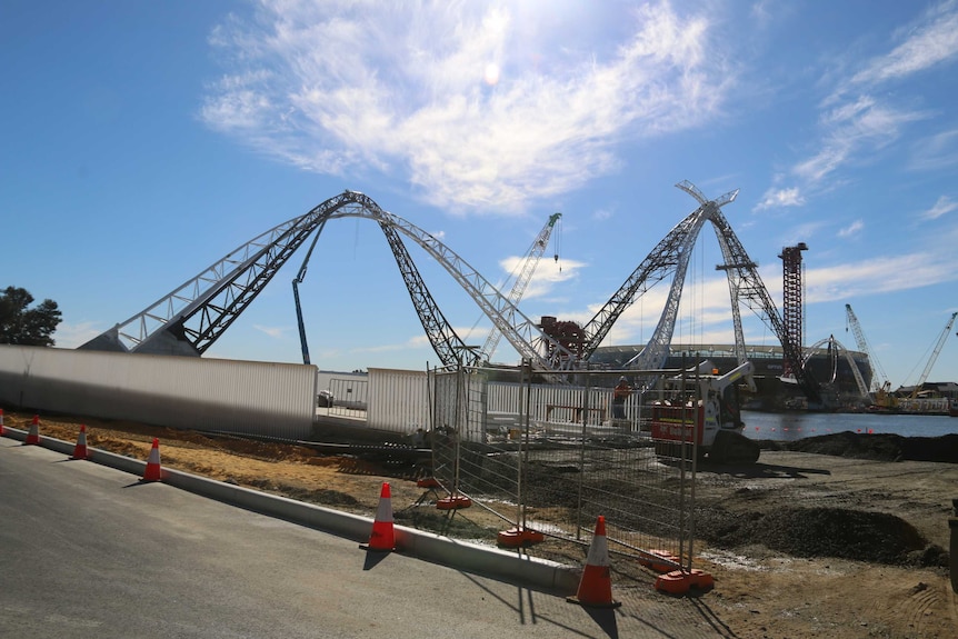 A fence outside the construction site at the Perth Stadium pedestrian bridge with arches and the stadium in the background.