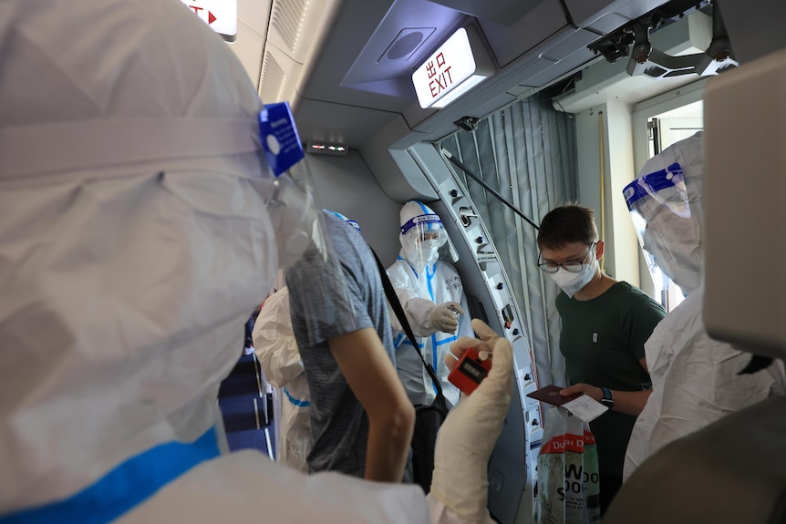 A man walks onto a plane holding a passport while several people in full hazmat suits look on