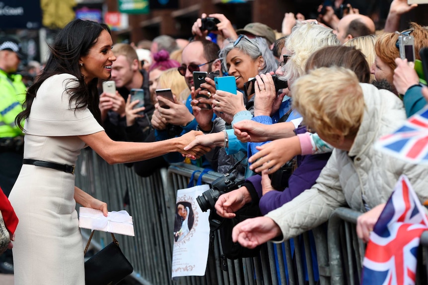 Meghan, Duchess of Sussex shakes hands with a fan as she walks along a street.