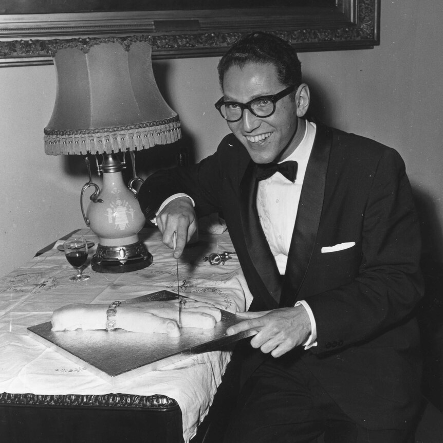 Tom Lehrer, wearing a tuxedo, cuts a cake in the shape of a human hand.