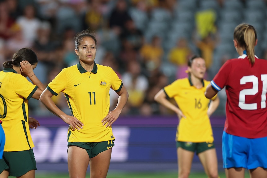 A soccer player wearing yellow and green puts her hands on her hips during a game with team-mates in the background