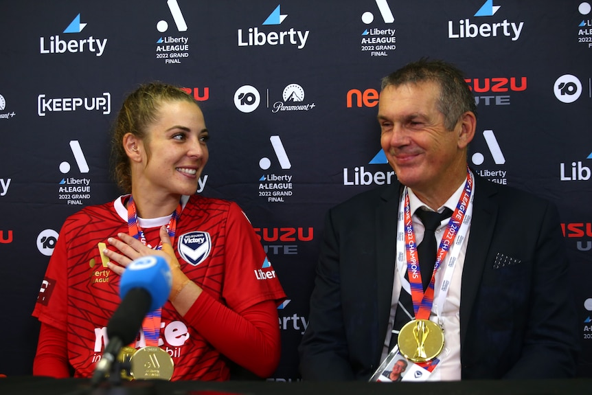 A soccer player wearing red and a man in a jacket sit at a media desk wearing medals