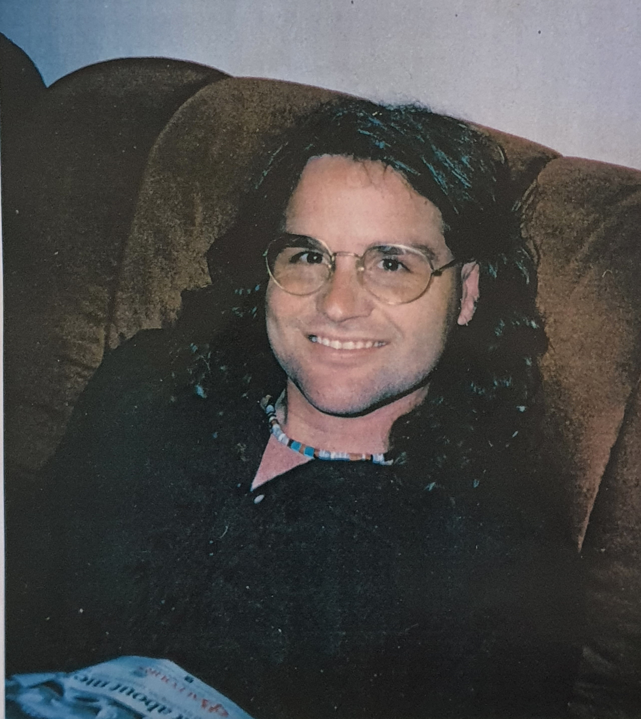A man with glasses and long, dark, curly hair smiles as he sits on a couch.