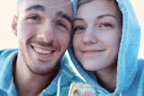 A young bald man and young woman with blonde hair wear grey hoodies, smiling.