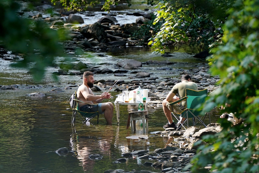 Two men sit in a river having a barbecue. 