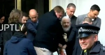 Julian Assange is held by officials. His beard is long, his hair white, as he is led out of embassy.