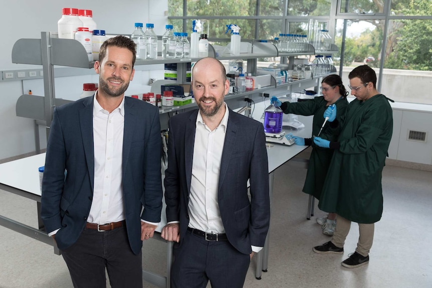 Two men in suits smiling at the camera with a man and woman working in a science lab in the background.