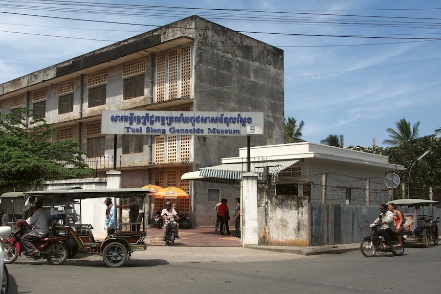 A building surrounded with barbed wire pictured from the street with tuk tuks and motorbikes on the road.