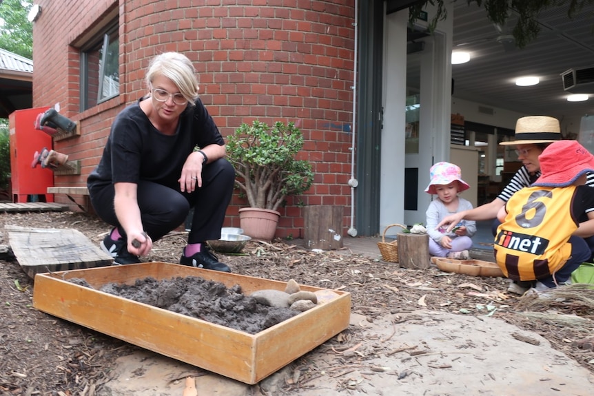 Nicole Messer crouches down and handles dirt in a box, as young children play nearby.