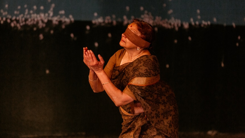 A Sri Lankan woman in her mid 60s wearing a blindfold and brown sari, she kneels with hands up