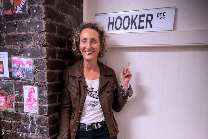 Candi Forrest smiles as she stands in an office with the street sign Hooker Parade and pink painted sign 'Sex work is work'.