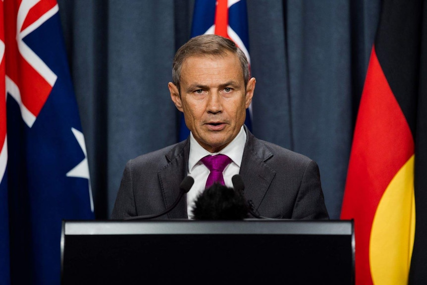 Roger Cook speaks at a podium with Australian, WA and the Aboriginal flag in the background.