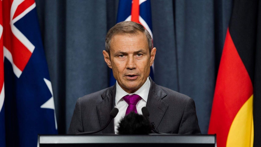 Roger Cook speaks at a podium with Australian, WA and the Aboriginal flag in the background.