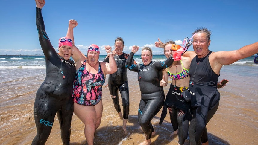 A group of six women in bathers standing on a beach, cheering.