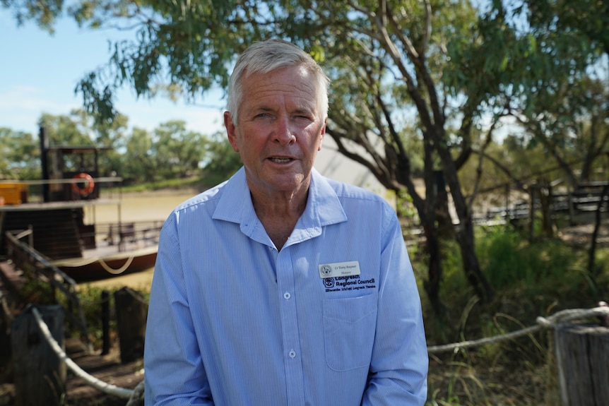  A man stands facing the camera with a boat in the background