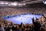 A picture of a tennis match under lights on a court surrounded by a packed crowd in Melbourne.
