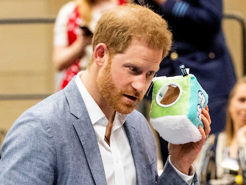 Prince Harry holds a small plush cube up to his ear, apparently listening for a rattle.