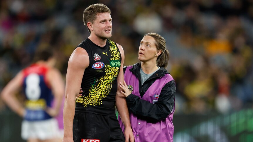 A Richmond player looks emotional as he stands on the ground while a medic puts her hand on his arm.