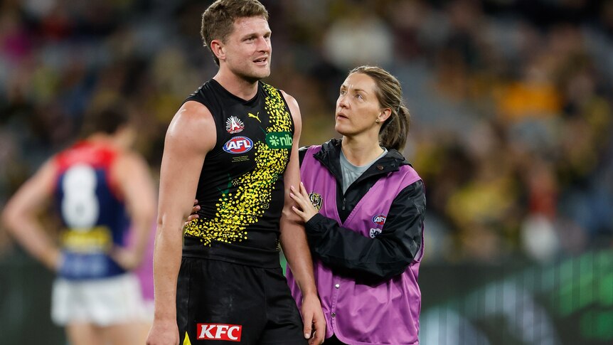 A Richmond player looks emotional as he stands on the ground while a medic puts her hand on his arm.