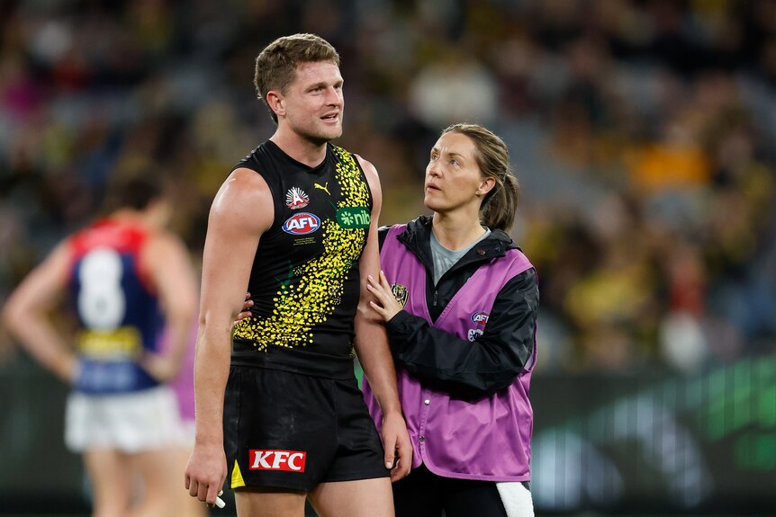 A Richmond player looks emotional as he stands on the ground while a medic puts her hand on his arm.