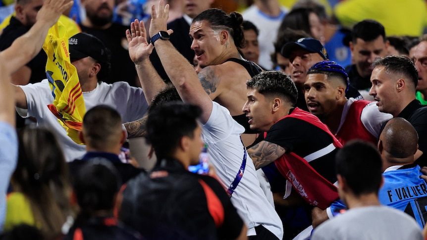 A Uruguayan footballer in a black singlet advances with hands raised towards Colombian fans in the stands after a game.