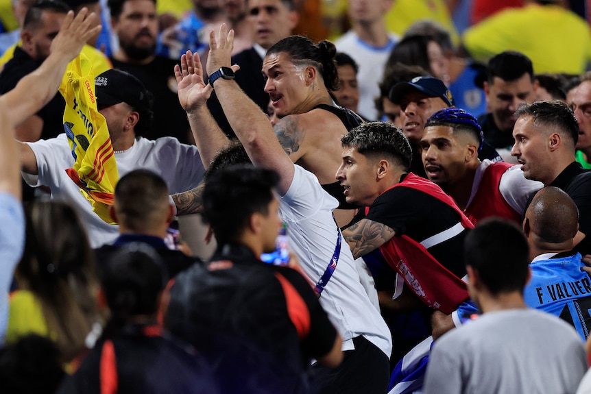 A Uruguayan footballer in a black singlet advances with hands raised towards Colombian fans in the stands after a game.