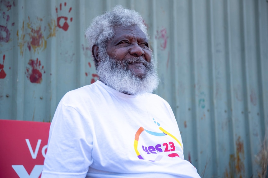 An elderly Indigenous man wearing a Yes23 shirt in front of a remote building