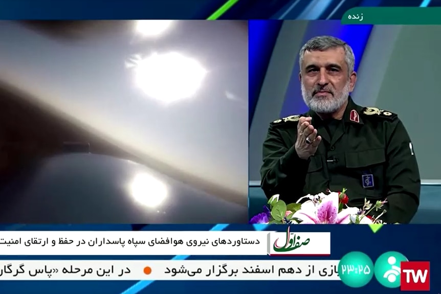 A grey-haired man in a military uniform sits in a TV studio as a split-screen view shows a missile in mid-air.