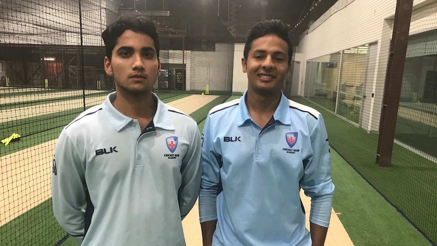 Two young cricketers stand smiling in some indoor nets.