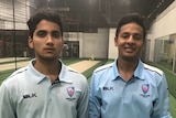 Two young cricketers stand smiling in some indoor nets.