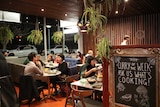 People sit at tables and on couches in the cafe, a sign in the foreground reads some recipes.