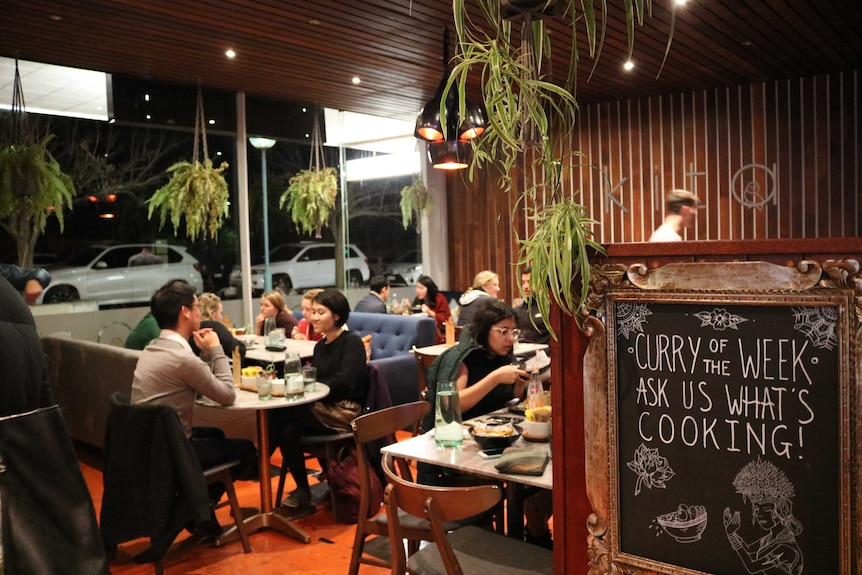 People sit at tables and on couches in the cafe, a sign in the foreground reads some recipes.