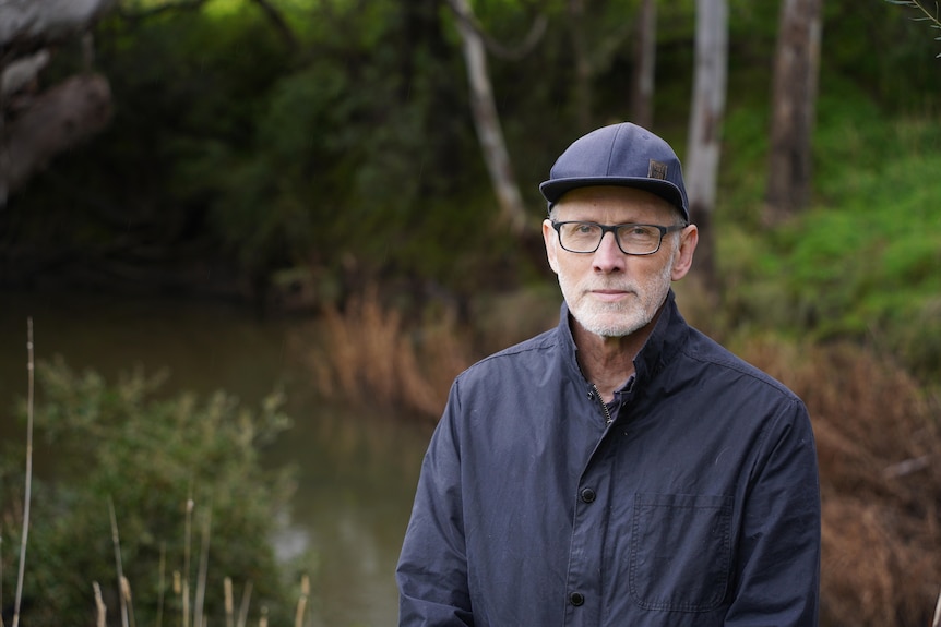 a man with glasses and a short beard in a blue jacket and a blue cap stands before a river bank