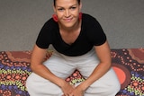 A young woman sits cross legged, smiling on top of a mat featuring an Aboriginal design.