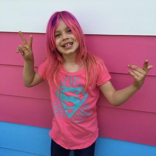 A young girl with pink hair stands in front of a wall painted pink, blue and white