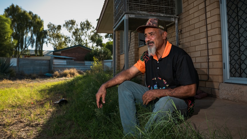 Nigel, a man weraing black Tshirt with orange collar, and a cap, sitting in his backyard.