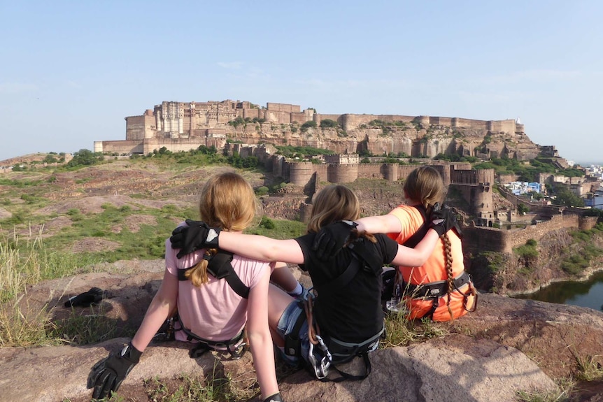 Three girls, with their arms around each other, look out at a castle.