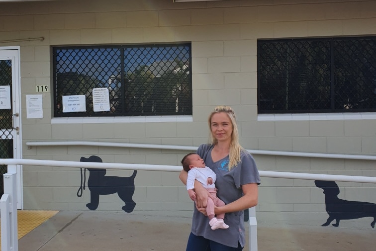 woman holding small baby stand outside vet clinic with a grey and blue sign reading gold city vet clinic