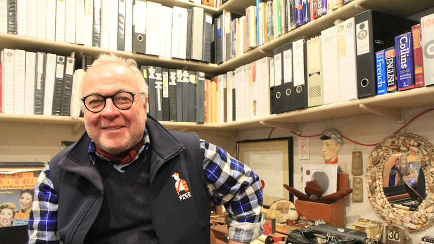 A man smiling in front of a desk with a typewriter, and a heap of folders.