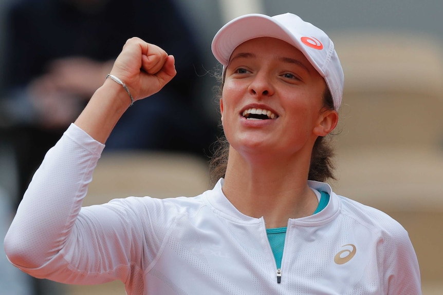 Iga Swiatek, wearing all white and an all white cap raises her fist in triumph at Roland Garros.