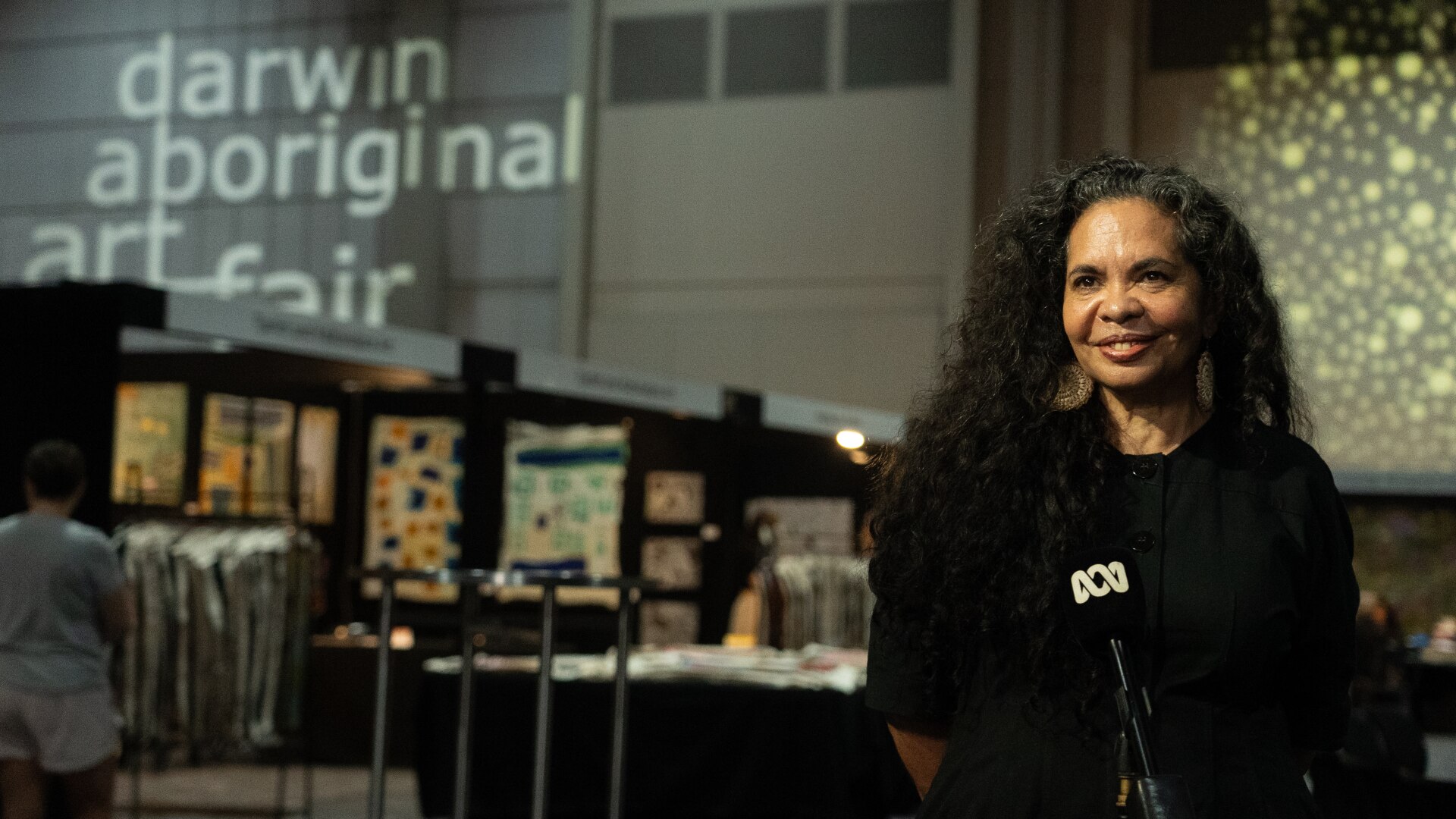 A smiling woman sitting and speaking into a microphone as an art fair takes place in the background.