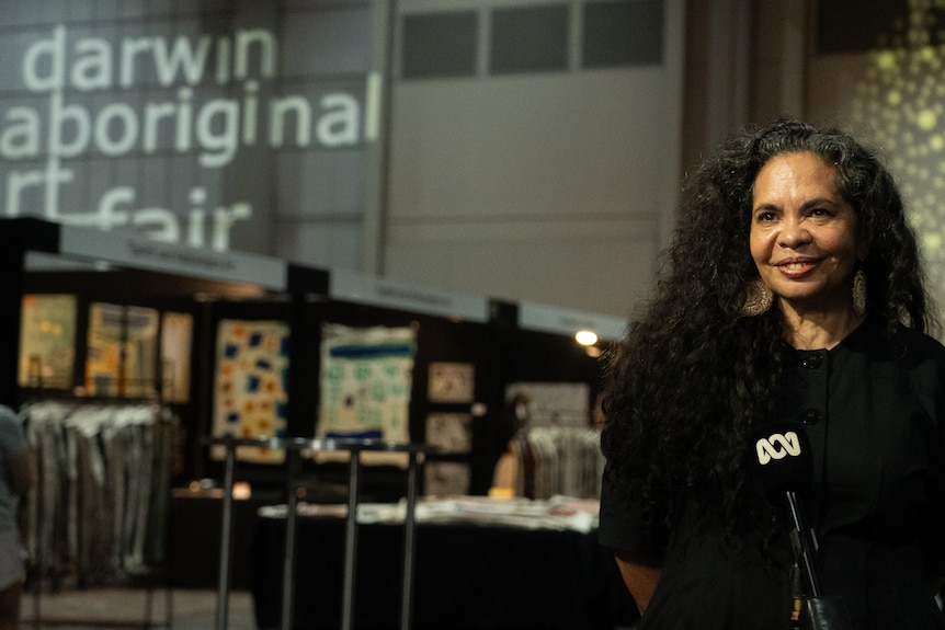 A smiling woman sitting and speaking into a microphone as an art fair takes place in the background.